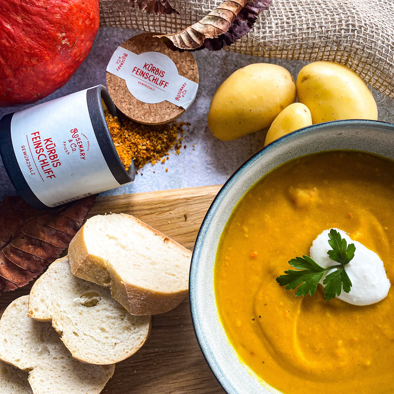 Eine Schüssel Kürbissuppe mit Sahne und Petersilie, daneben Gewürz in einer Pappdose, Kartoffeln und Brotscheiben.
