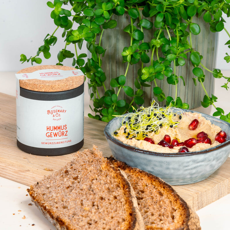 Pappdose mit Hummus-Gewürz, eine Schale Hummus mit Granatapfelkernen und Sprossen, Brot und grüne Pflanze im Hintergrund.