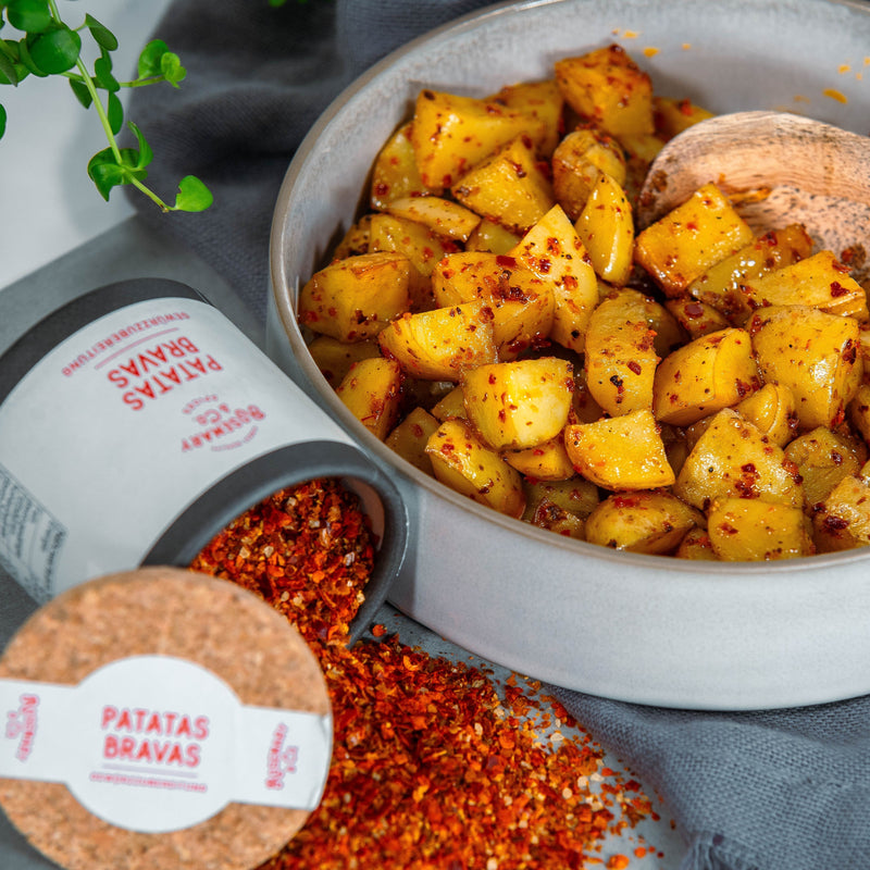 Graue Schüssel mit gewürfelten Kartoffeln, daneben eine umgestürzte Pappdose mit Patatas-Bravas-Gewürz und Löffel.