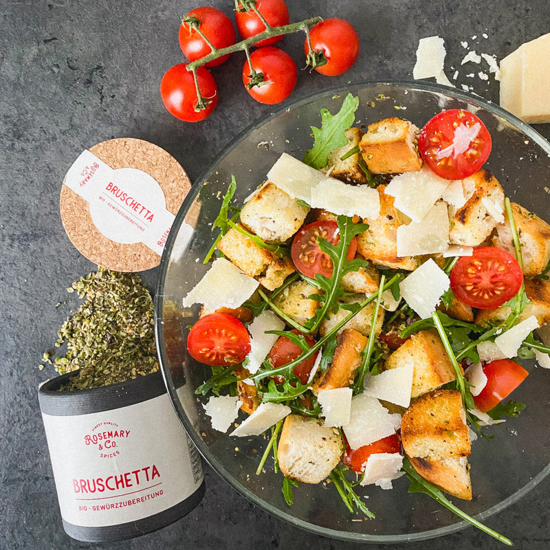 Salat mit Tomaten, Rucola, Croutons und Parmesan in einer Glasschüssel, daneben Gewürze und frische Tomaten.