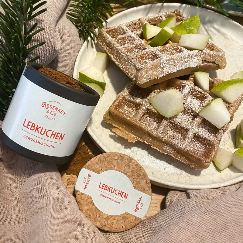 Dose mit Lebkuchengewürz neben einem Teller mit Waffeln und Apfelstücken, garniert mit Puderzucker.