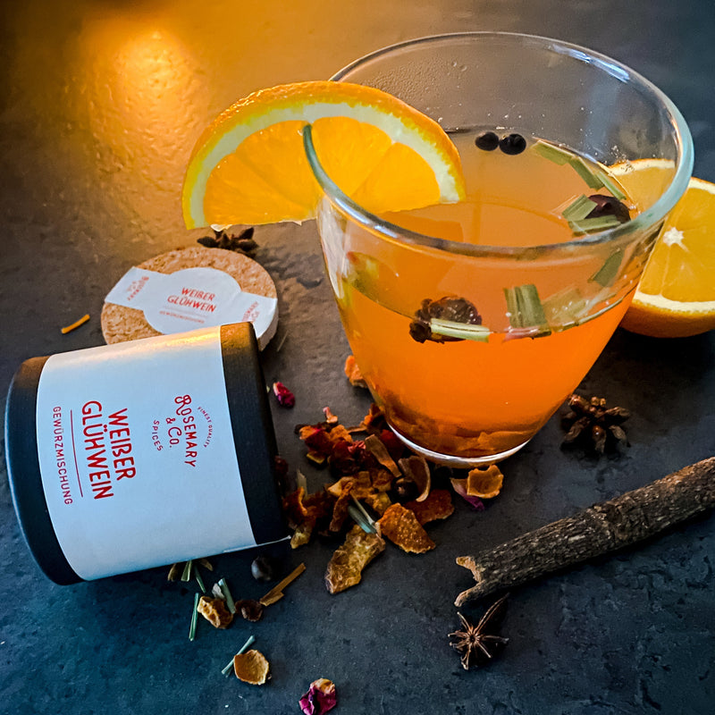 Glas mit weißem Glühwein und Orangenscheibe, daneben eine Dose Gewürzmischung und getrocknete Zutaten.