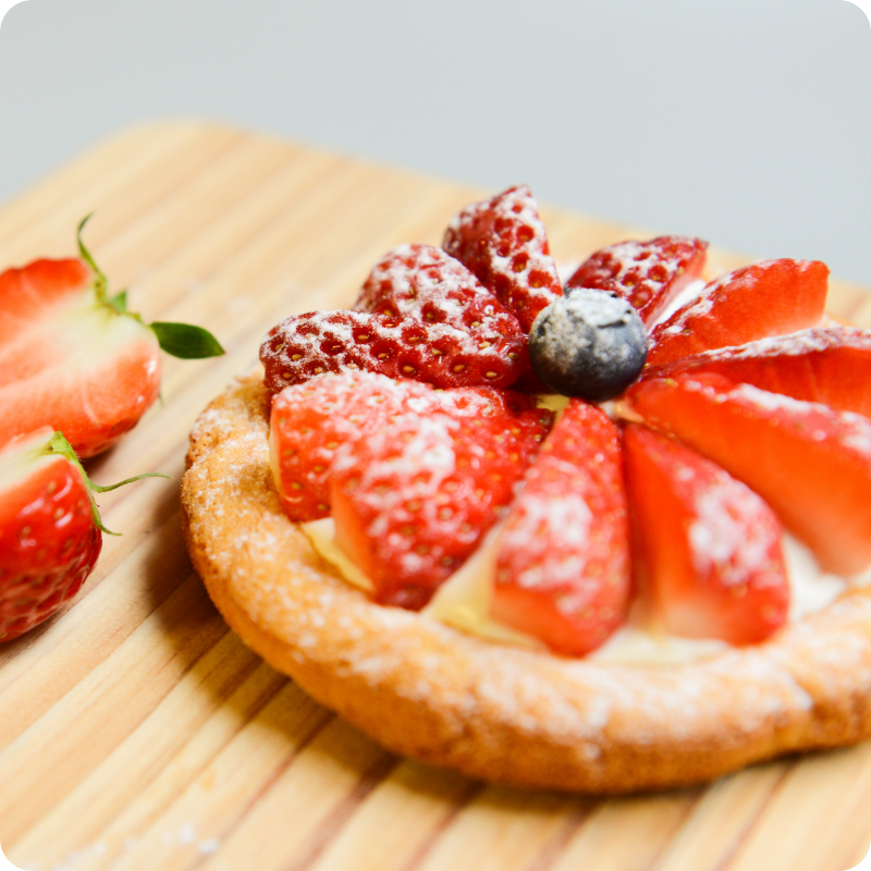 Erdbeertörtchen mit geschnittenen Erdbeeren und einer Heidelbeere, bestäubt mit Puderzucker, auf einem Holzbrett.