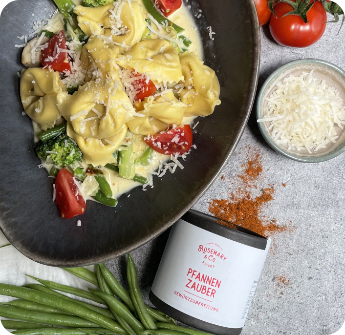 Tortellini mit Brokkoli und Tomaten in einer Schale, daneben grüne Bohnen, Gewürzdose und geriebener Käse.