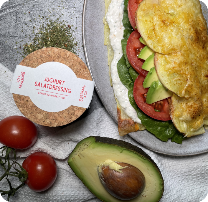 Ein Teller mit Omelett, Tomaten, Avocado und Joghurt-Dressing neben frischen Tomaten und einer Avocado.