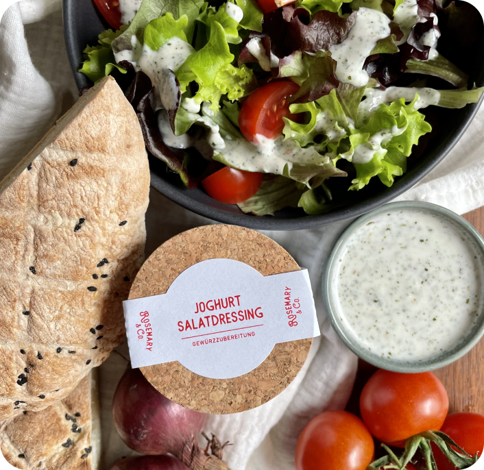 Schüssel mit Salat und Dressing, Pappdose mit Joghurt-Salatdressing, Baguette, Tomaten und Zwiebeln auf Leinentuch.