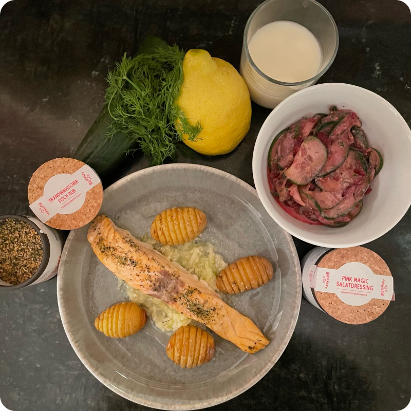 Gegrillter Lachs auf einem Teller mit Kartoffeln, daneben eine Pappdose, Dill, Zitrone, Gurkensalat und ein Glas Milch.