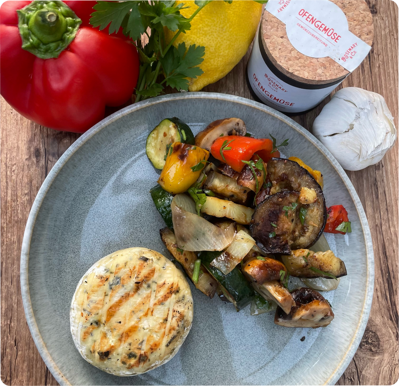 Gegrillter Käse und Ofengemüse auf grauem Teller, mit Paprika, Zitrone, Knoblauch und Dose auf Holzuntergrund.