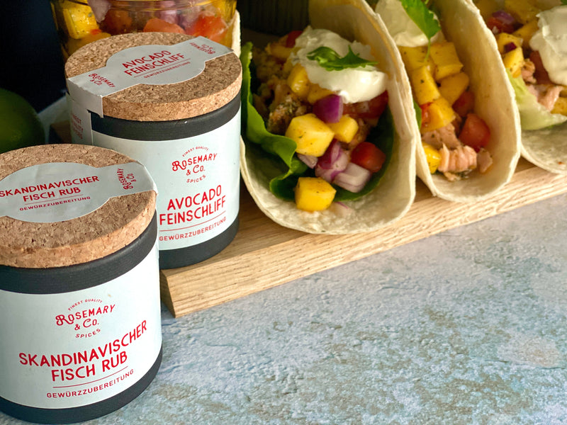 Zwei Pappdosen mit Gewürzen neben gefüllten Tacos auf einem Holzbrett.