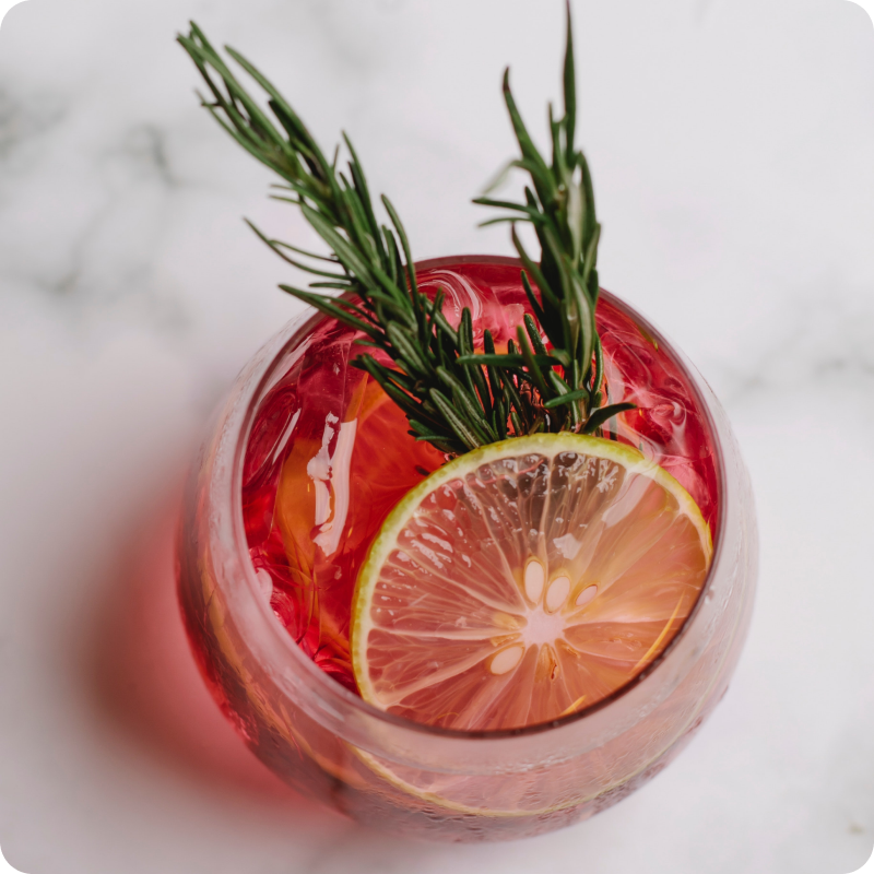Ein Glas mit rotem Getränk, Eiswürfeln, einer Zitronenscheibe und einem Rosmarinzweig auf einem Marmortisch.