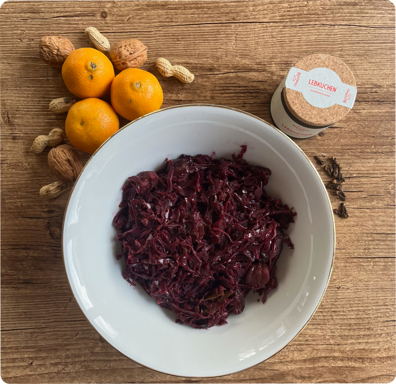 Eine Schale mit Rotkohl, daneben Mandarinen, Nüsse und eine Dose Lebkuchen auf einem Holztisch.