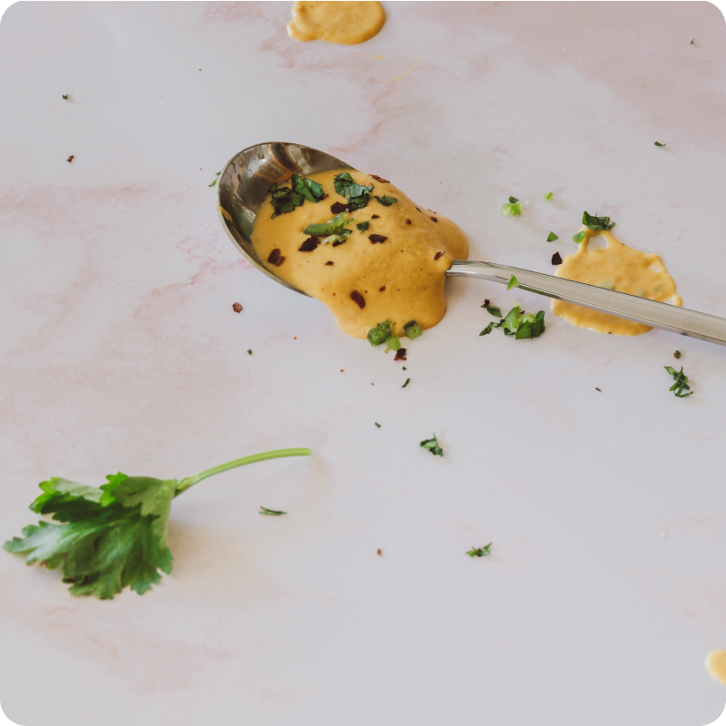 Ein Löffel mit gelber Sauce, garniert mit Kräutern, liegt auf einer marmorierten Oberfläche neben einem Petersilienblatt.