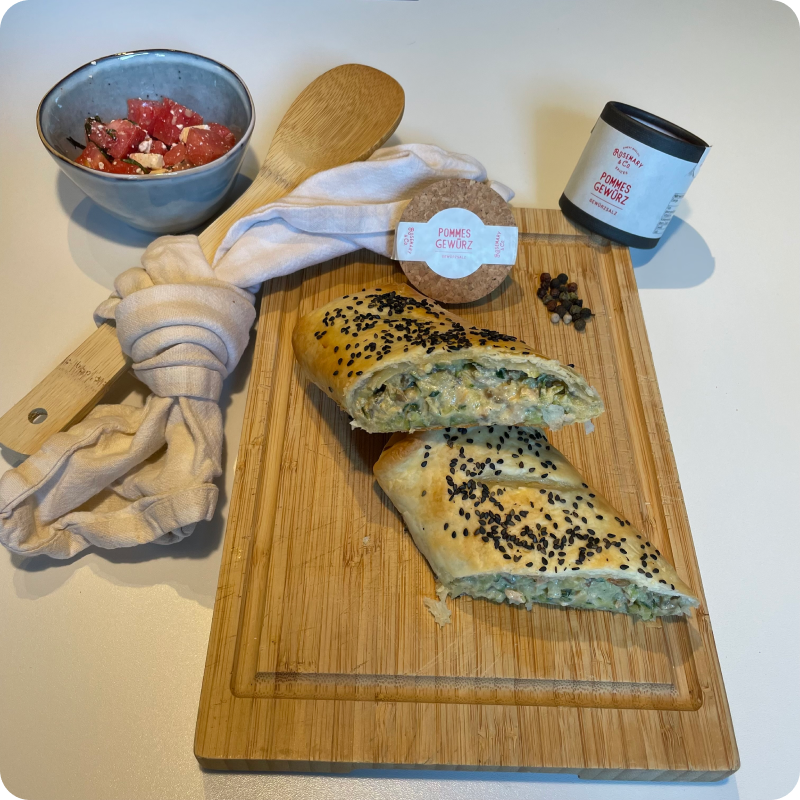 Geschnittenes Gebäck mit schwarzem Sesam auf einem Holzbrett, daneben eine Schale Salat und eine Dose Gewürz.