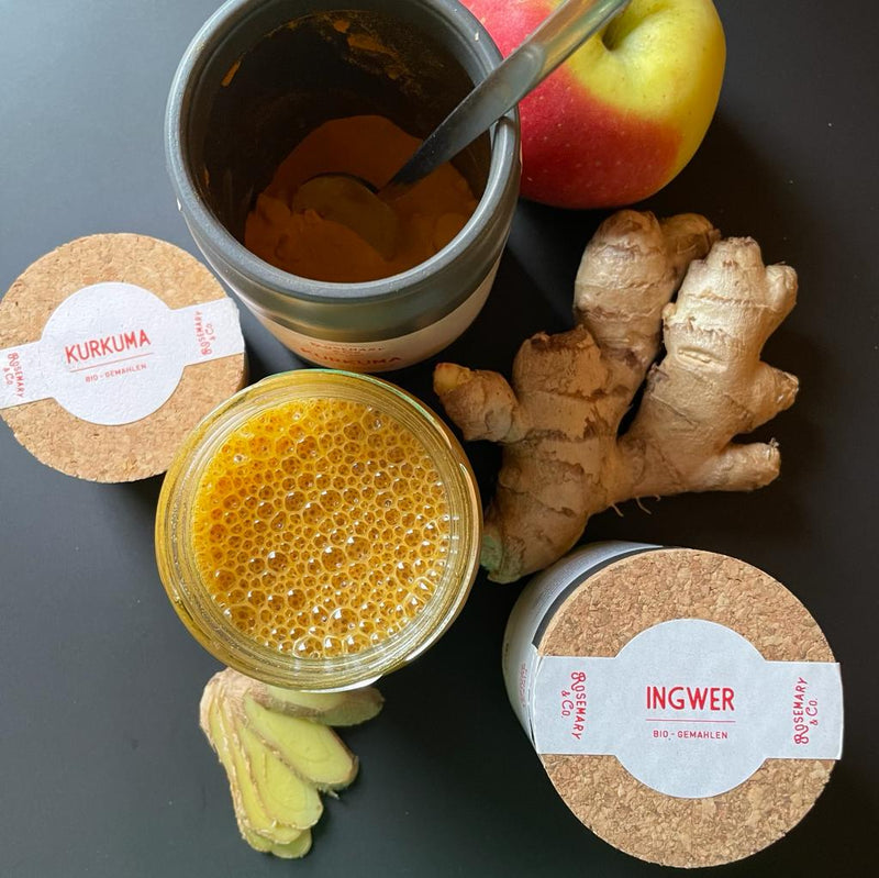 Zutaten auf einem Tisch: Eine Kurkuma-Dose, Ingwer, Apfel und ein Glas mit schäumendem Getränk.