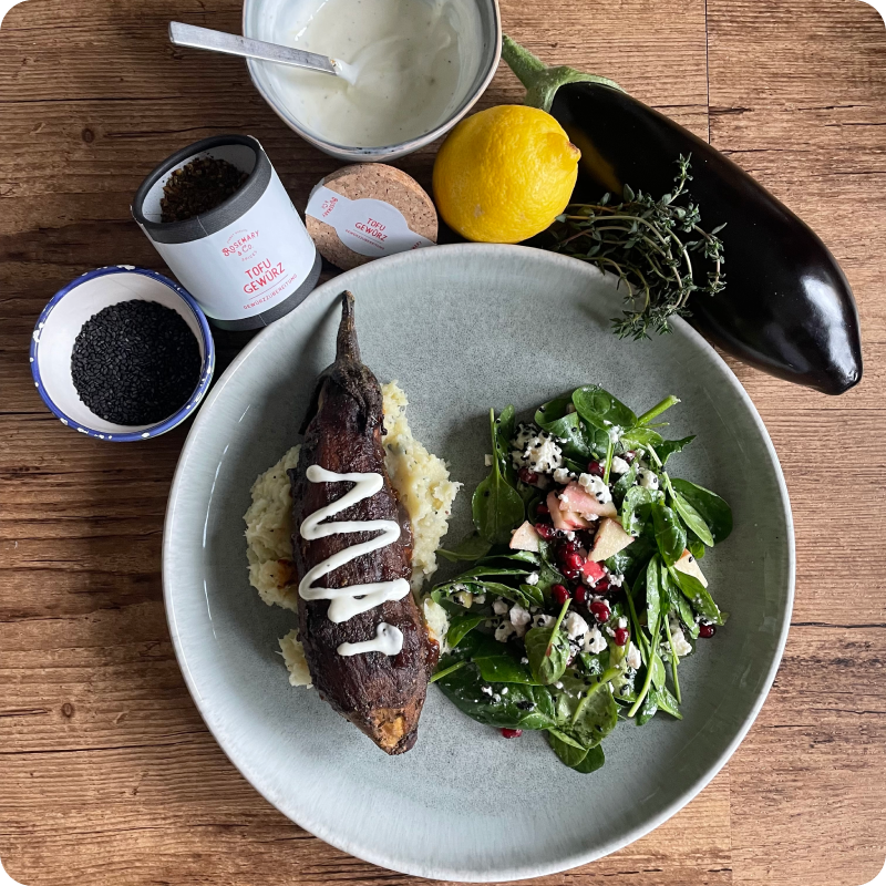 Ein Teller mit gefüllter Aubergine auf Kartoffelpüree und Salat. Daneben Zutaten wie Gewürze, Zitrone und Aubergine.