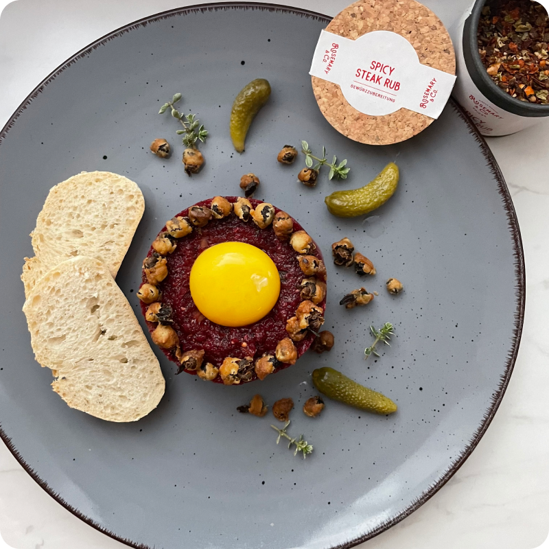 Grauer Teller mit Tatar, rohem Eigelb, Gewürzen, eingelegten Gurken, Brot und einer Gewürzdose.