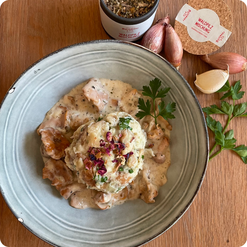 Ein Teller mit Knödel in Pilzrahmsauce, dekoriert mit Petersilie und Blüten, daneben Zutaten und eine Pappdose.