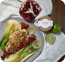Ein Teller mit gebratenem Hähnchen, Granatapfel, Limettenscheiben und Pak Choi, daneben eine Limette und eine Pappdose.