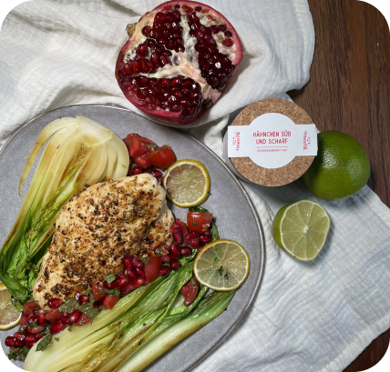 Ein Teller mit gebratenem Hähnchen, Granatapfel, Limettenscheiben und Pak Choi, daneben eine Limette und eine Pappdose.