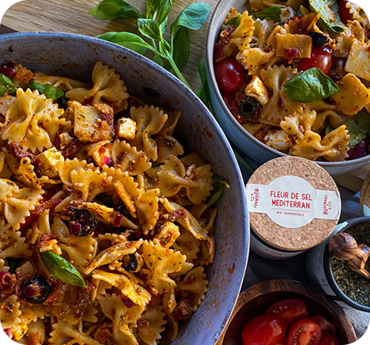 Zwei Schüsseln mit Pasta-Salat, dekoriert mit Basilikumblättern, neben einer Pappdose mit der Aufschrift "Fleur de Sel Mediterran".