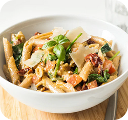 Schüssel mit Penne-Pasta, frischem Basilikum, Tomaten, Spinat und Parmesanscheiben auf einem Holzbrett.