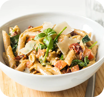 Schüssel mit Penne-Pasta, frischem Basilikum, Tomaten, Spinat und Parmesanscheiben auf einem Holzbrett.