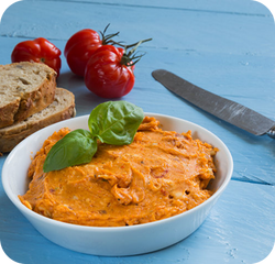 Rötliche Kräuterbutte, dahinter Brot, Tomaten und ein Messer zu sehen.