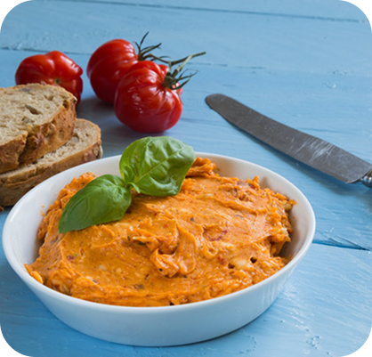 Rötliche Kräuterbutte, dahinter Brot, Tomaten und ein Messer zu sehen.