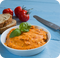Rötliche Kräuterbutte, dahinter Brot, Tomaten und ein Messer zu sehen.