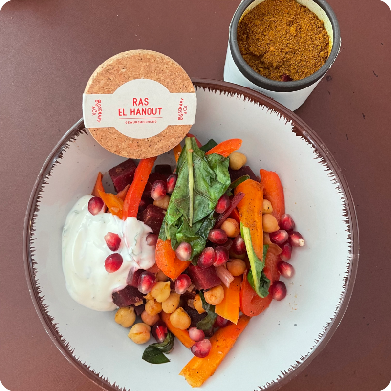 Weiße Schale mit buntem Salat, Kichererbsen, Granatapfelkernen und Soße, daneben Gewürzdose mit Ras El Hanout.