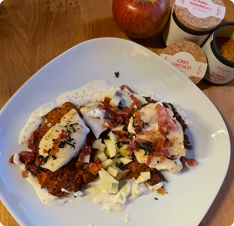 Ein Teller mit verziertem Essen, geräuchertem Fisch, Speck und Apfelwürfeln, daneben Gewürzdosen und ein Apfel.