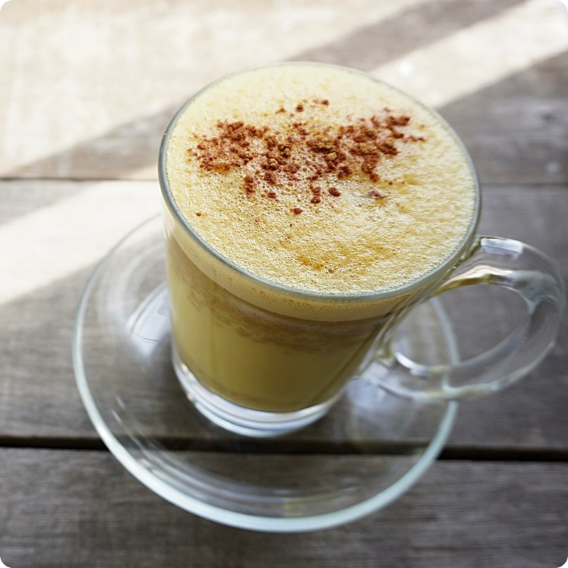 Ein Glas Latte mit Schaum und Zimt auf einem hölzernen Tisch bei Tageslicht.