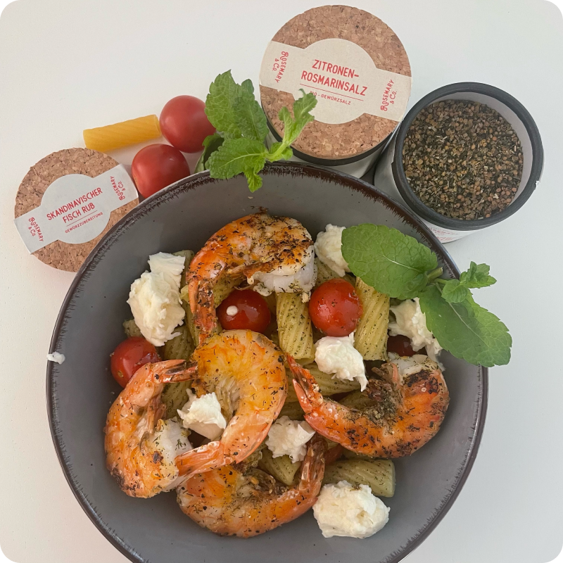 Schwarze Schüssel mit Garnelen, Tomaten, Nudeln und Käse, umgeben von Gewürzdosen und Minzblättern.
