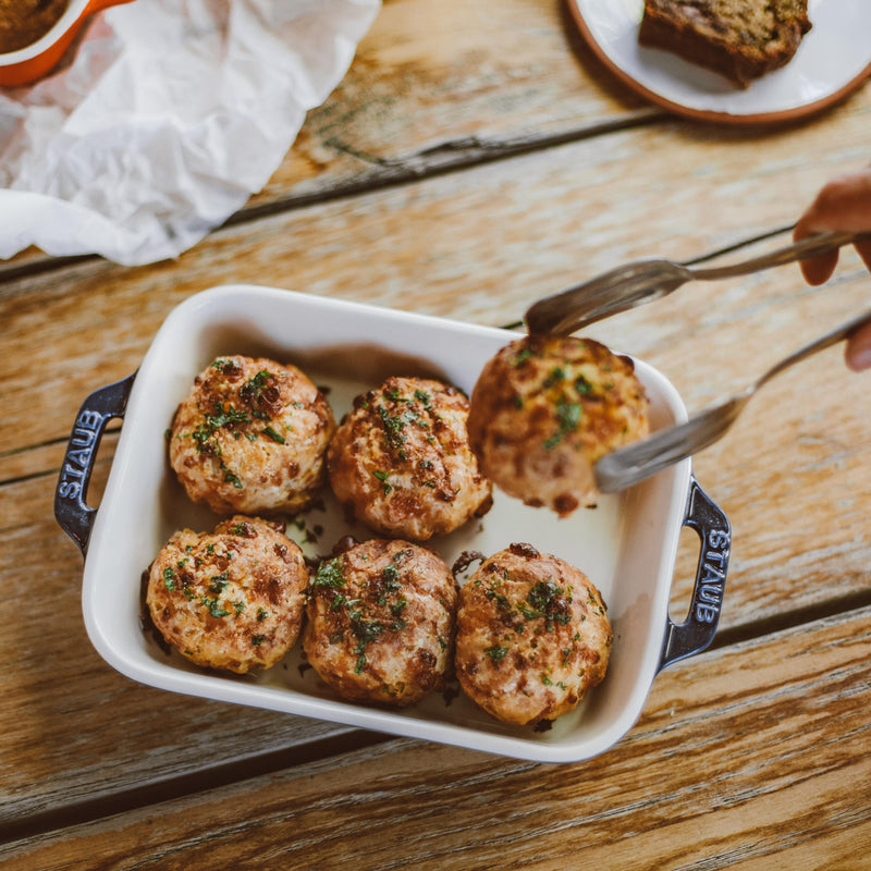 Sechs gebackene Fleischbällchen in einer weißen Auflaufform auf einem Holztisch, eines wird mit der Zange serviert.