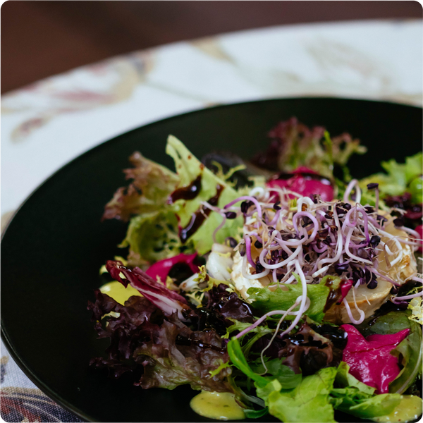 Frischer bunter Salat mit Sprossen, Rotkohl und Dressing auf einem schwarzen Teller, auf einem Tisch mit Musterdecke.