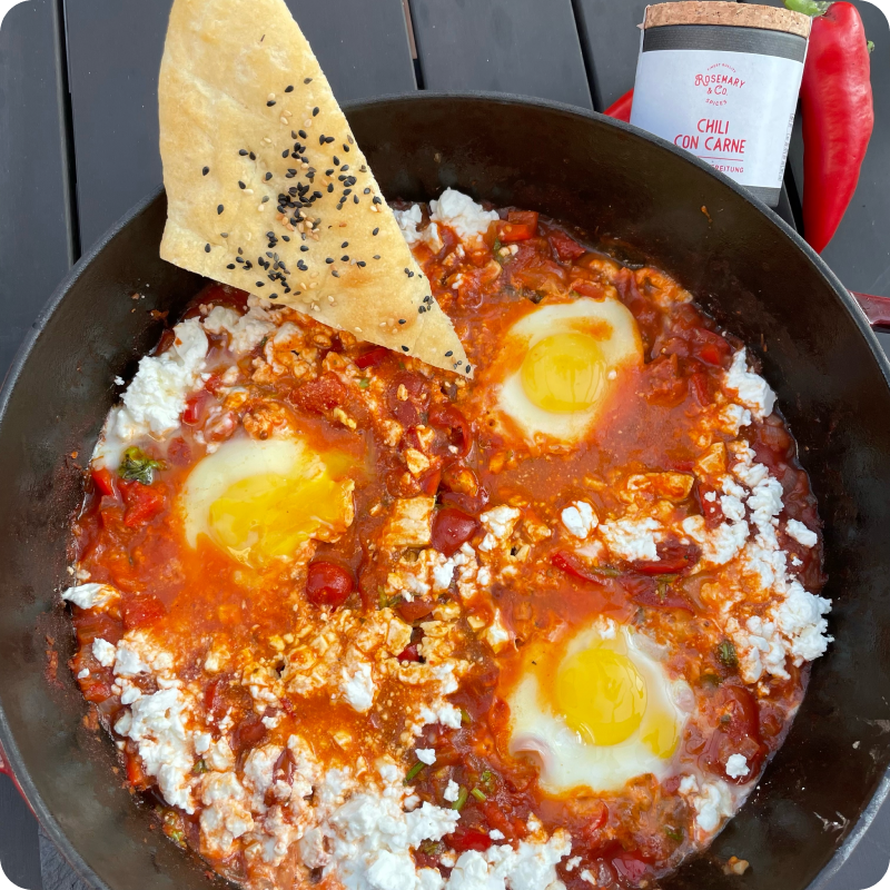 Pfanne mit Shakshuka, drei Eiern, und Sesamfladenbrot, daneben eine Gewürzdose für Chili con Carne.