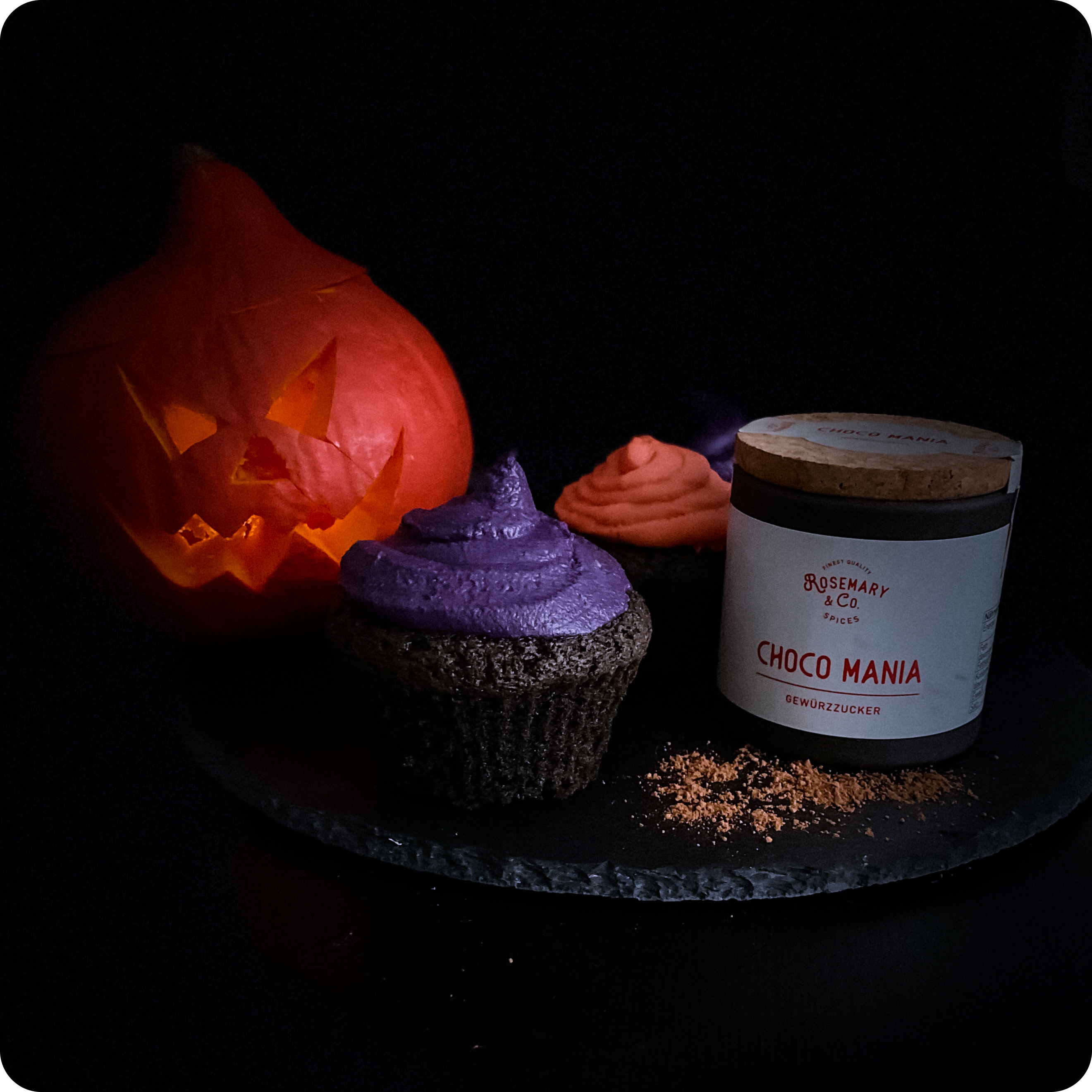 Schnitzkürbis, zwei Cupcakes mit lila Zuckerguss, Dose CHOCO MANIA von Rosemary & Co., dunkle Unterlage