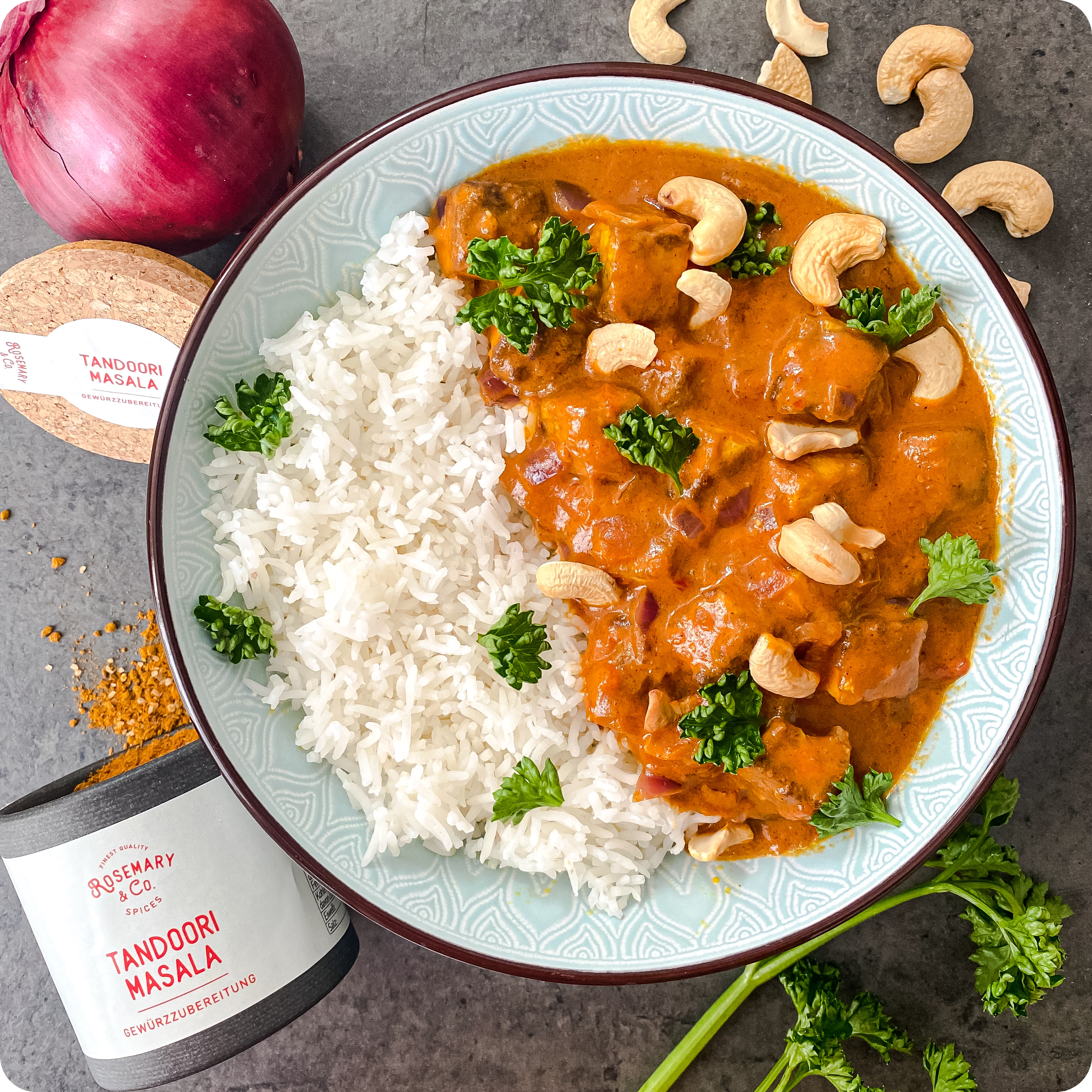 Teller mit Reis, Tandoori-Curry, Cashews und Petersilie, daneben eine Pappdose Tandoori Masala und eine rote Zwiebel.