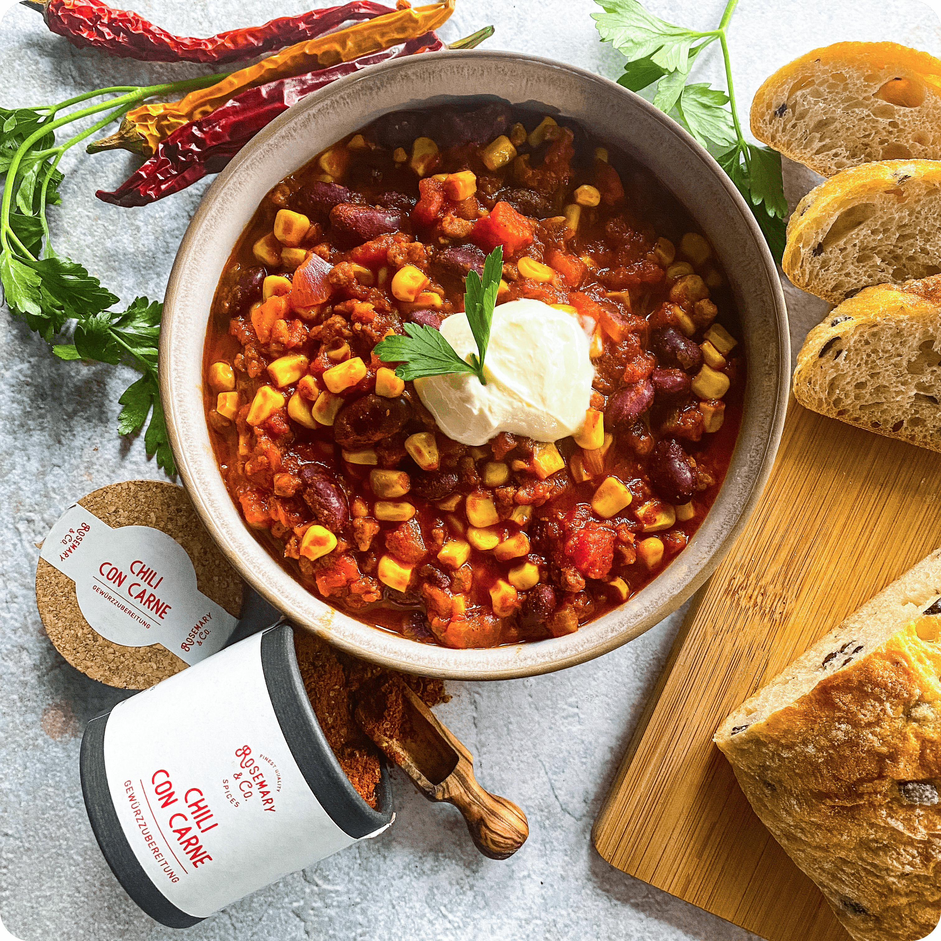 Dosis Rosemary & Co Gewürze neben Chili con Carne mit Mais, Bohnen, Sauerrahm, Brot und frischen Kräutern