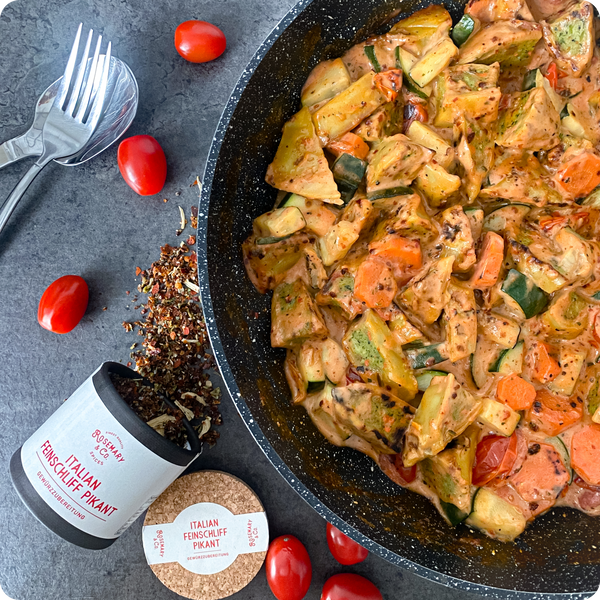 Eine Pfanne mit vegetarischer Pasta, umgeben von Tomaten, Gewürzstreu und einer Pappdose für italienische Gewürze.