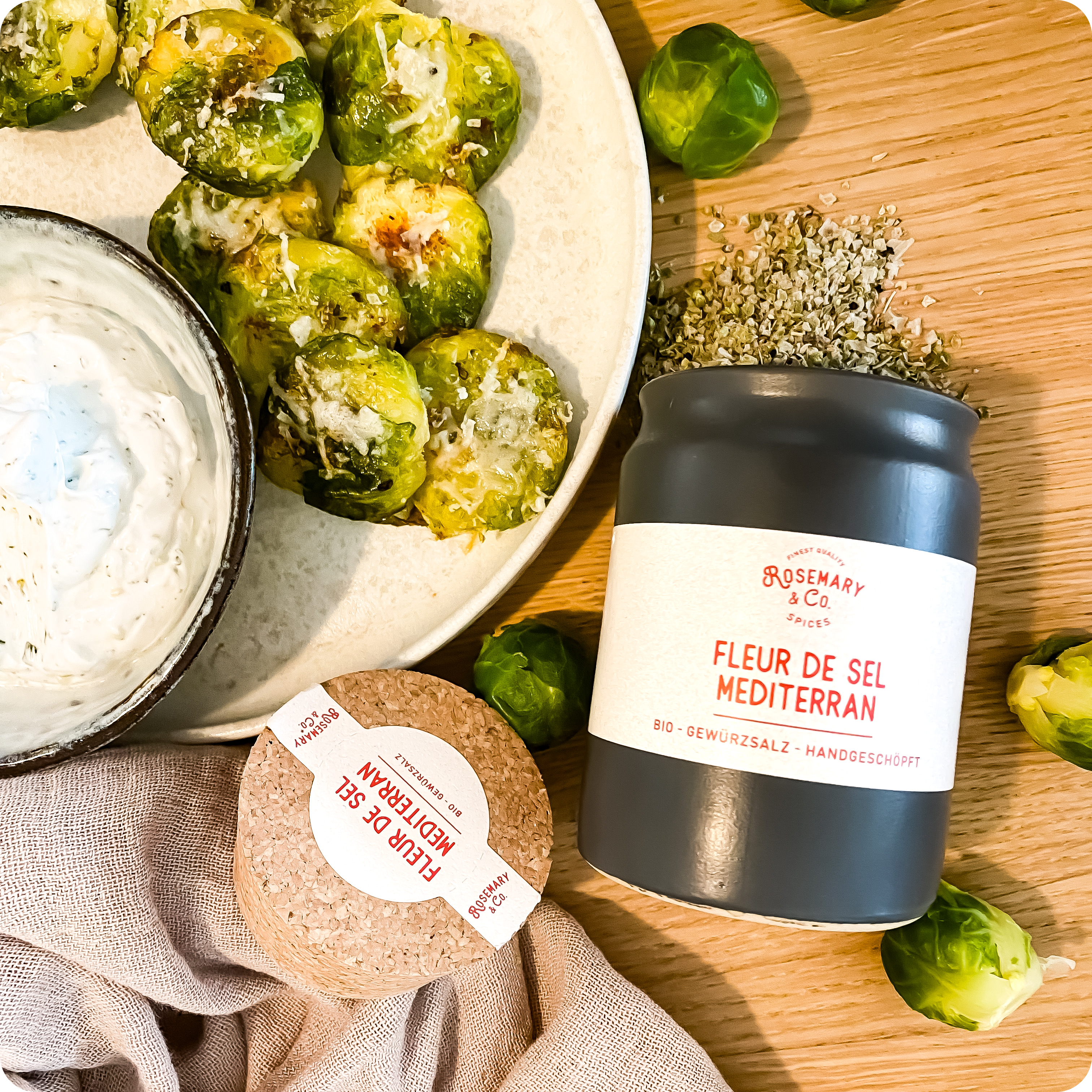 Ein Teller mit Rosenkohl und Dip neben einer Dose Fleur de Sel Mediterran auf einem Holztisch.