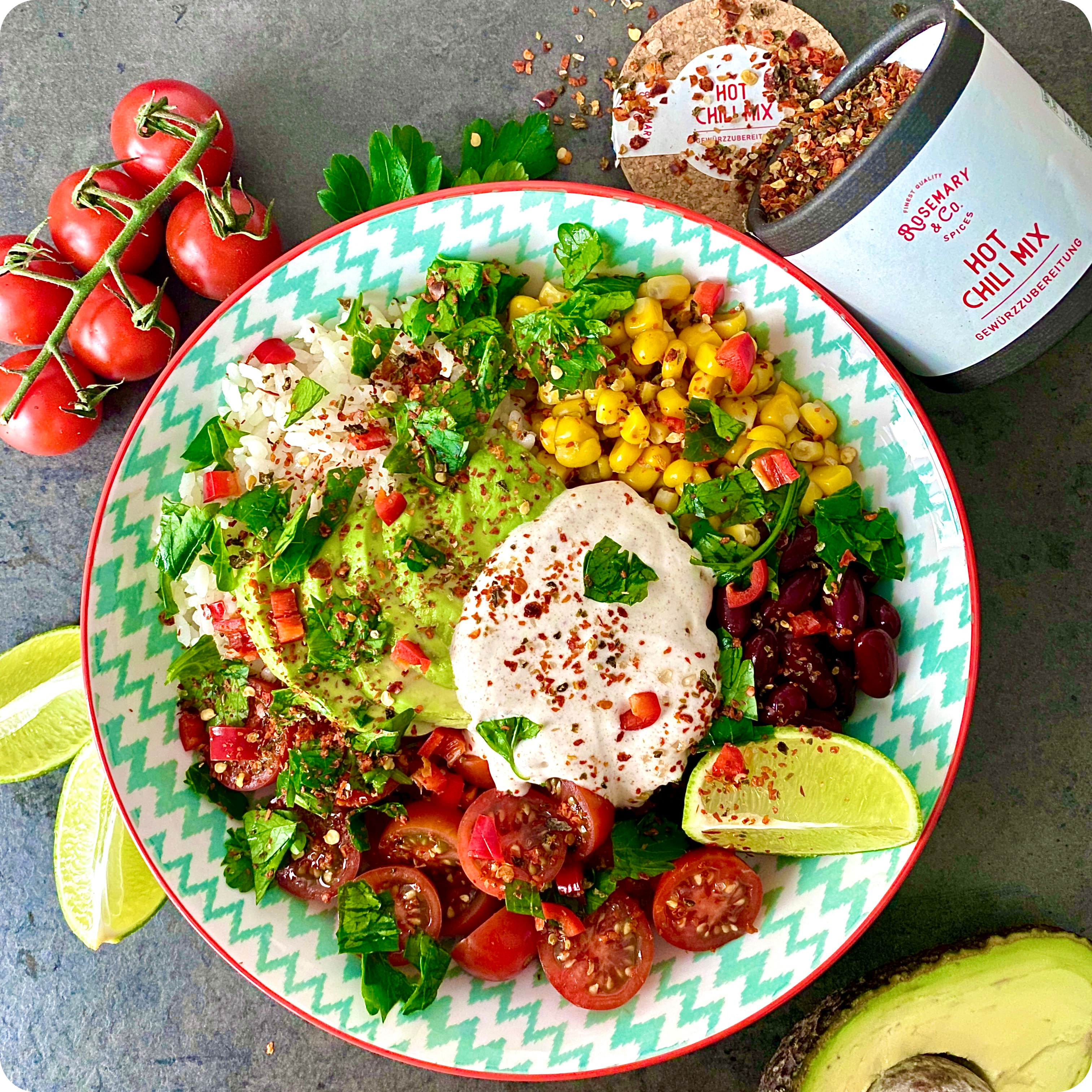 Eine farbenfrohe Schüssel mit Reis, Mais, Kidneybohnen, Kirschtomaten, Guacamole, Sahnesauce und gehackten Kräutern ist mit dem Gewürz-Hot-Chili-Mix von Rosemary & Co. garniert. Limettenspalten, eine Avocado und der Chili-Mix-Behälter stehen daneben auf der grauen Oberfläche.